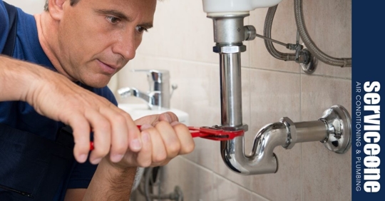 A middle aged caucasian man holding a red pipe wrench with two hands and using it to twist a silver pipe under a sink