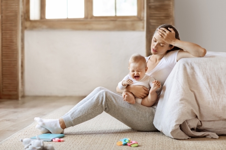 Young woman, hand on her head, sitting on the floor with a crying baby