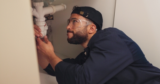 attractive young plumber wearing goggles and tightening pipe under sink