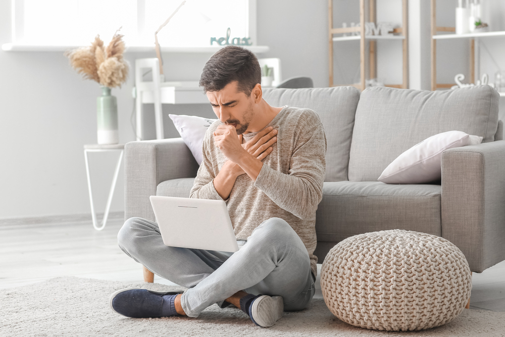 young man sitting crosslegged on the floor with his computer coughing