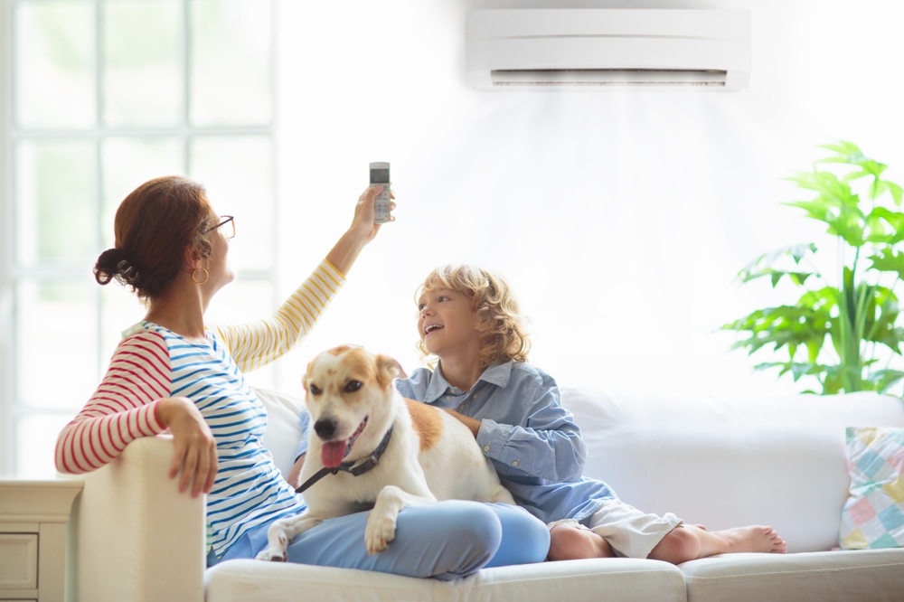 mom, dog, and young child sit on a couch laughing while she aims the AC remote control