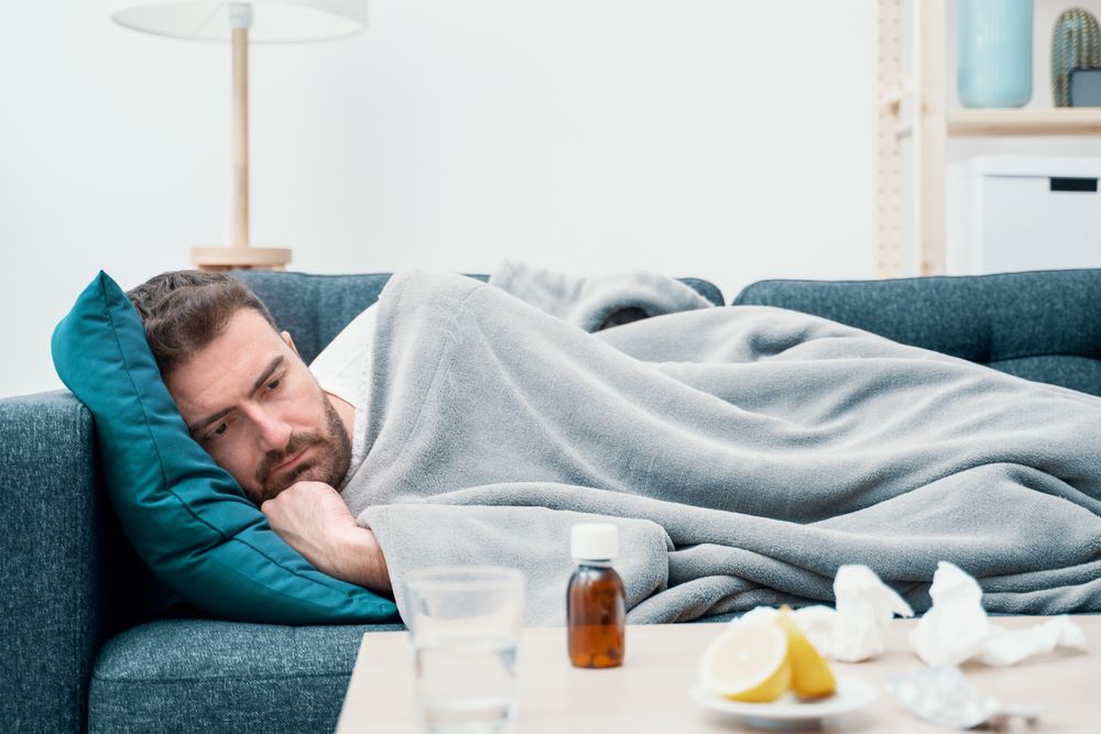 man on couch, covered with blanket  with tissues and tea and lemon nearby